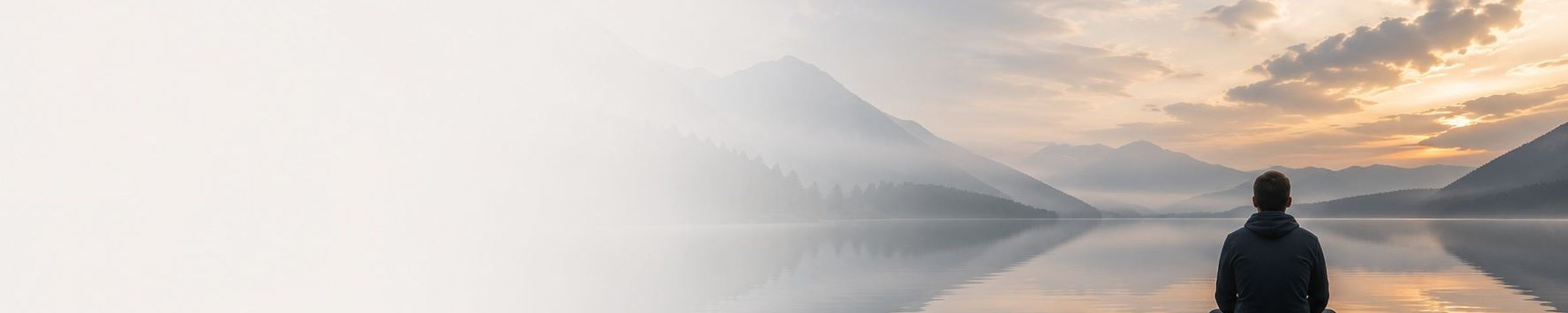 Iemand zit alleen aan een spiegelglad bergmeer bij zonsopkomst; de bergen weerspiegelen in het water.